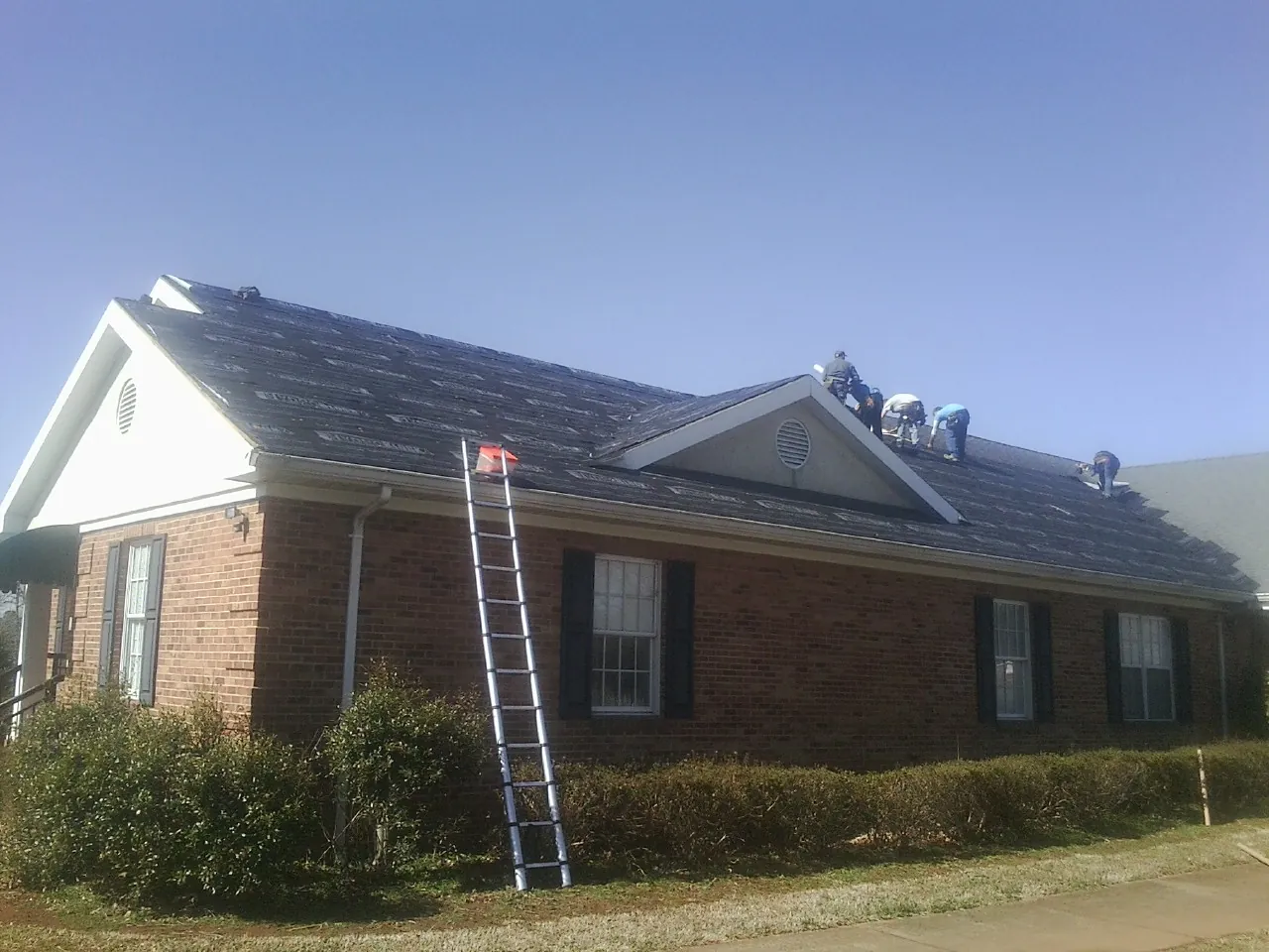 Crew tearing shingles off a brick ranch roof with a ladder set — Metal Roof Repair work by our crew in Vida