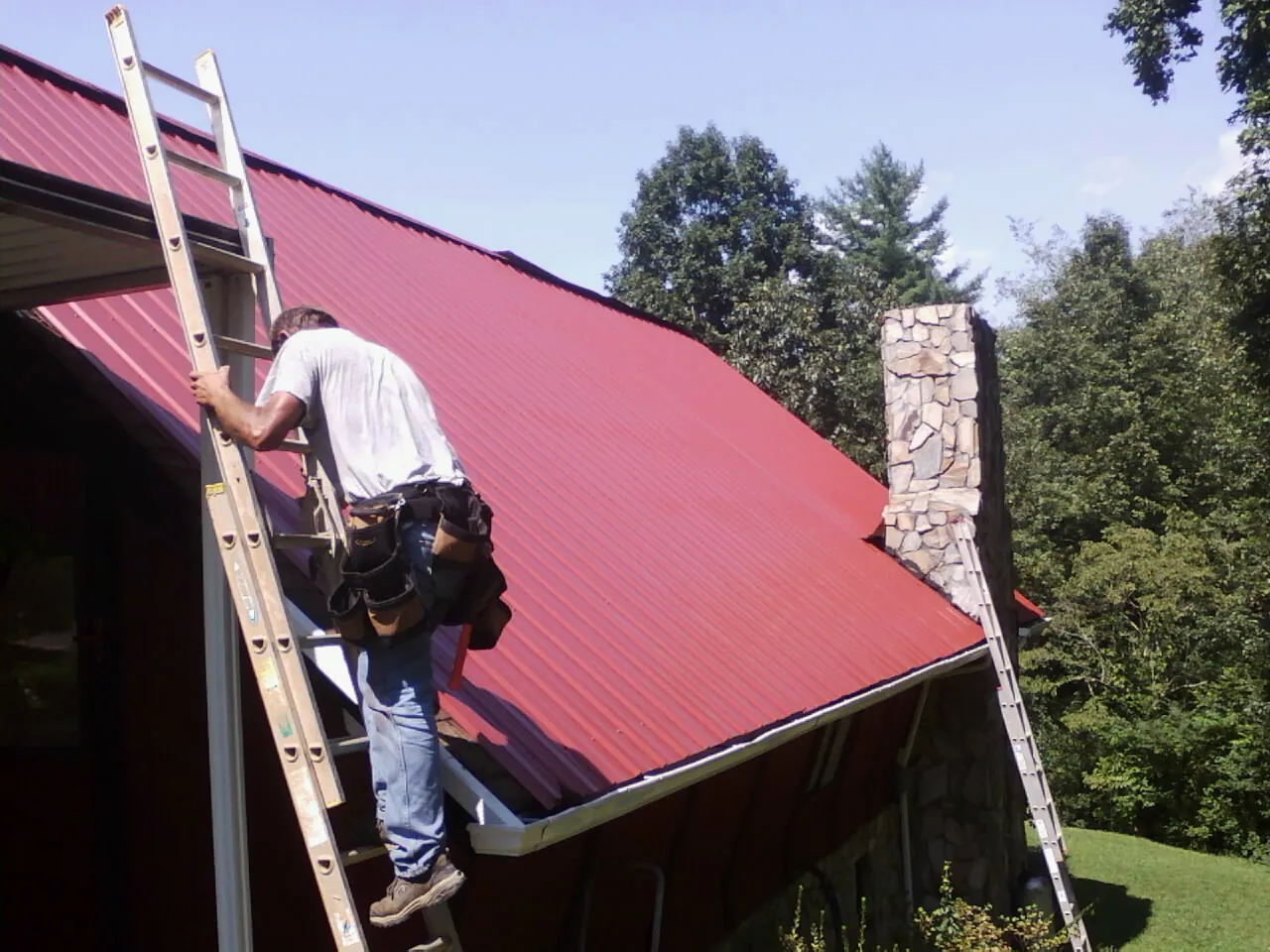 Roofer on a ladder beside a red metal roof and stone chimney — Soffit & Fascia Repair by Roofing Brothers in Vida